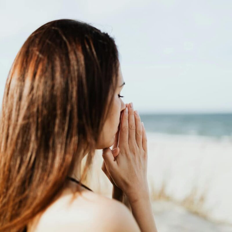 Woman at a beach looks out over the distance and holds hands to face in grief for her loss