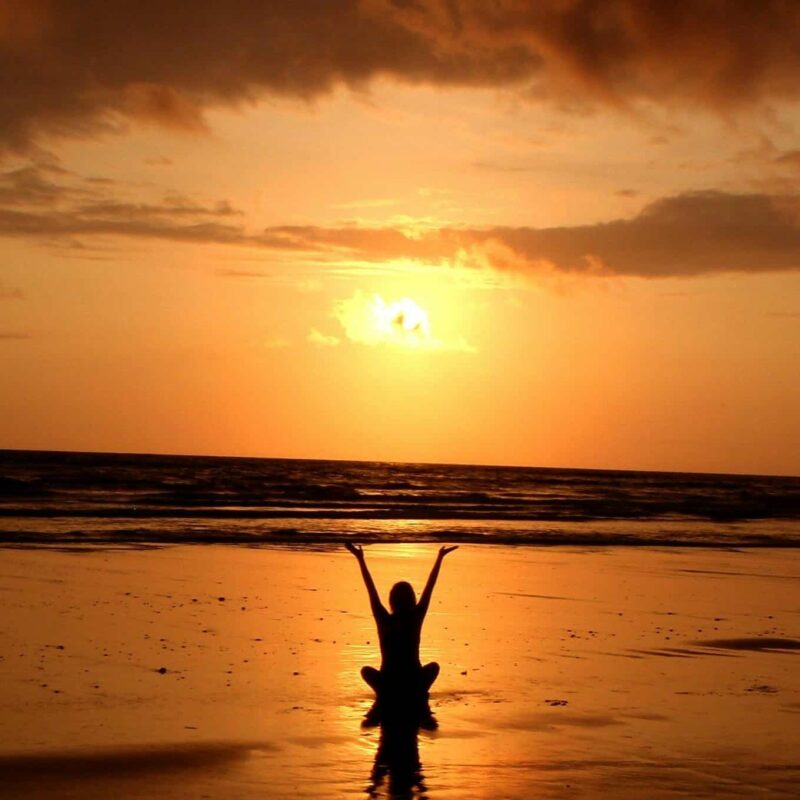 Woman at a beach at sunset sitting in the shallows with arms raised; she is balanced from her wellness treatment